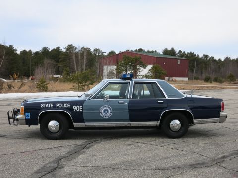 1991 Ford LTD Crown Victoria - Driver-side profile of a navy-and-gray Massachusetts State Police Ford cruiser showing the state-seal door decal and STATE POLICE 90E text.