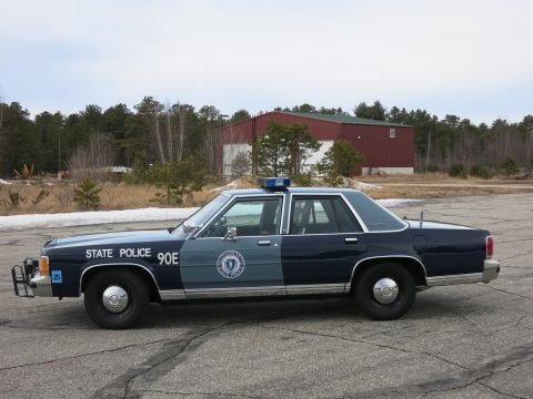 1991 Ford LTD Crown Victoria - Driver-side profile of a two-tone Massachusetts State Police Ford cruiser parked on cracked pavement with a red metal building behind.