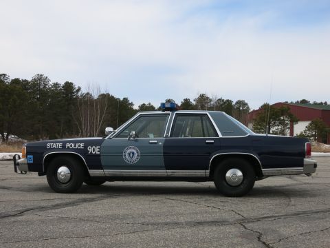 1991 Ford LTD Crown Victoria - Passenger-side profile of a two-tone navy and gray Massachusetts State Police Ford cruiser with state-seal decal and STATE POLICE 90E lette…