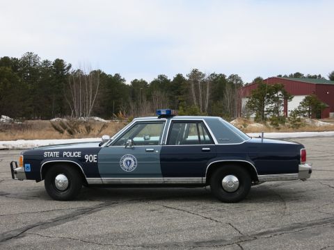 1991 Ford LTD Crown Victoria - Driver-side profile of a navy-and-gray Massachusetts State Police Ford cruiser parked on pavement before bare trees and a red building.