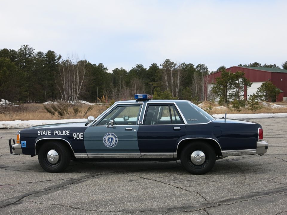 Massachusetts State Police Ford sedan, driver-side profile near tree line