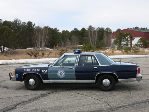 1991 Ford LTD Crown Victoria - Passenger-side profile of a two-tone Massachusetts State Police Ford cruiser with roof light bar and state-seal door decal.