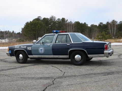 1991 Ford LTD Crown Victoria - Driver-side rear three-quarter view of a navy-and-gray Massachusetts State Police Ford cruiser on a snow-edged paved lot.