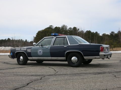 1991 Ford LTD Crown Victoria - Passenger-side rear three-quarter view of a two-tone Massachusetts State Police Ford cruiser parked on cracked pavement.