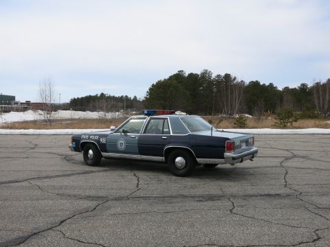 1991 Ford LTD Crown Victoria - two-tone Massachusetts State Police Ford cruiser viewed in rear three-quarter across a wide paved lot with snow piles behind.