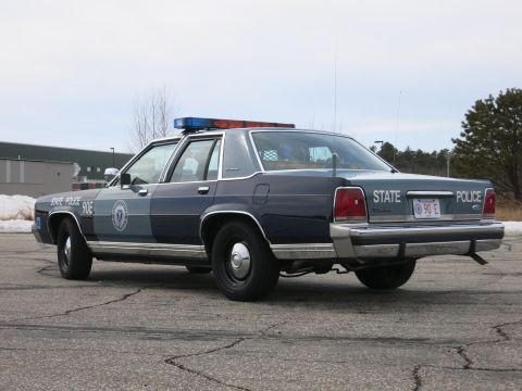 1991 Ford LTD Crown Victoria - Passenger-side rear three-quarter of a navy-and-gray Massachusetts State Police Ford cruiser with STATE POLICE 90E plate on cracked pavemen…