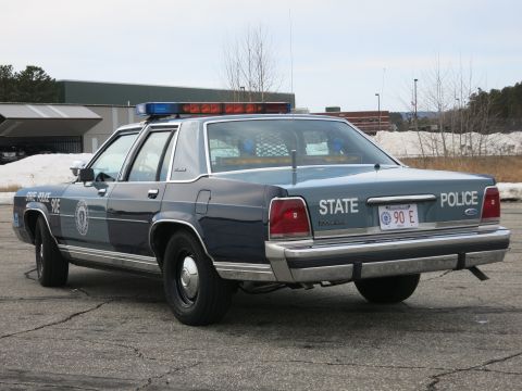1991 Ford LTD Crown Victoria - Rear three-quarter view of a navy-and-gray Massachusetts State Police cruiser with a Ford trunk badge and STATE POLICE 90E plate.