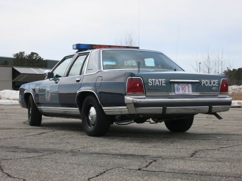 1991 Ford LTD Crown Victoria - Driver-side rear three-quarter view of a navy-and-gray Massachusetts State Police Ford cruiser with STATE POLICE 90E rear plate.