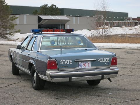1991 Ford LTD Crown Victoria - Rear of a two-tone blue Ford LTD Crown Victoria marked STATE POLICE with a roof light bar, parked on a paved lot near a warehouse.