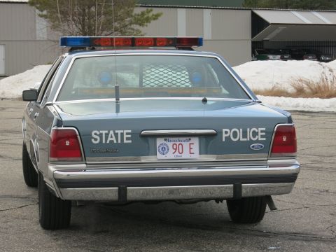 1991 Ford LTD Crown Victoria - Rear of a two-tone blue Ford LTD Crown Victoria State Police cruiser, red and blue roof light bar lit, parked outside a building.