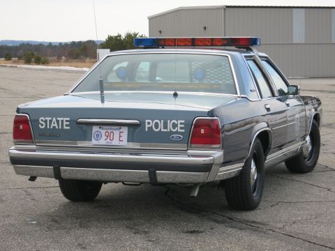 1991 Ford LTD Crown Victoria - Rear three-quarter view of a two-tone blue Ford LTD Crown Victoria State Police cruiser parked on a paved lot under a cloudy sky.