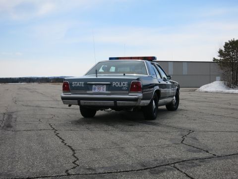 1991 Ford LTD Crown Victoria - Distant rear three-quarter view of a blue Ford State Police cruiser parked on a cracked tarmac lot.