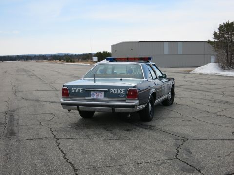 1991 Ford LTD Crown Victoria - Rear view of a blue Ford State Police cruiser parked at a distance on a cracked paved lot near a building.