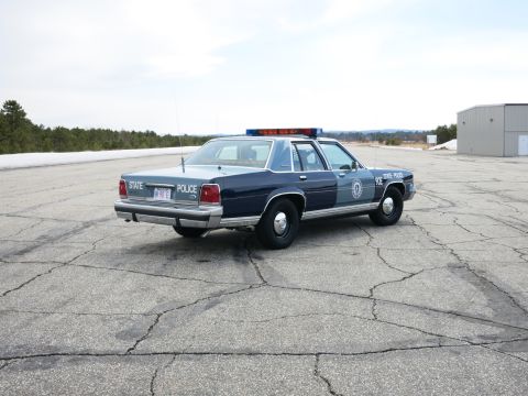 1991 Ford LTD Crown Victoria - Rear three-quarter view of a two-tone blue Ford State Police cruiser marked 90E STATE POLICE parked on a wide tarmac.