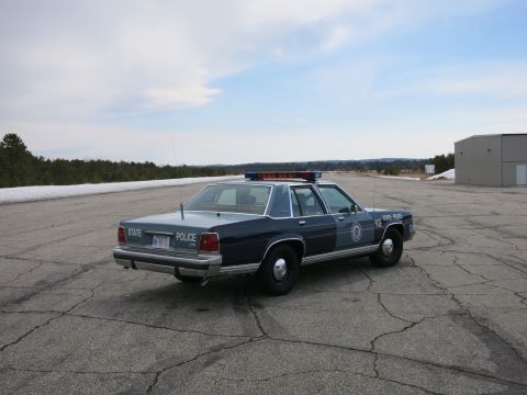 1991 Ford LTD Crown Victoria - Side-rear view of a two-tone blue Ford State Police cruiser marked 90E parked on a broad cracked lot.