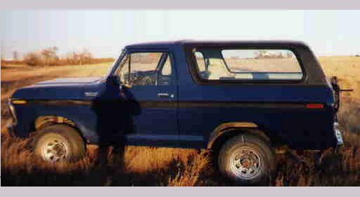 Dark blue two-door Ford Bronco SUV in profile in a grassy field with person by the open driver door