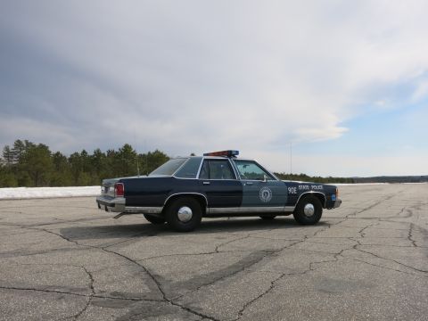 1991 Ford LTD Crown Victoria - Front-right side view of a two-tone blue Ford State Police cruiser marked 90E STATE POLICE parked on a wide cracked lot.