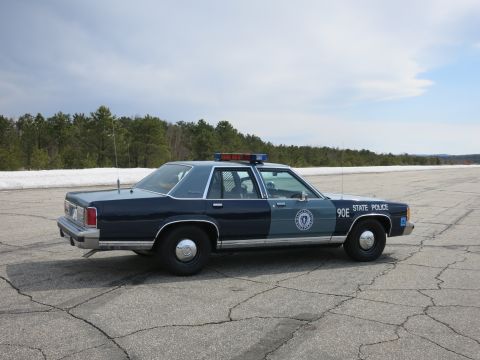 1991 Ford LTD Crown Victoria - two-tone blue Massachusetts State Police cruiser marked 90E sits in a paved lot with pine trees and snowbanks behind it.