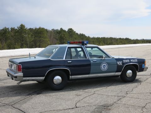 1991 Ford LTD Crown Victoria - blue Massachusetts State Police sedan marked 90E with a roof lightbar parked on cracked pavement near snowbanks.