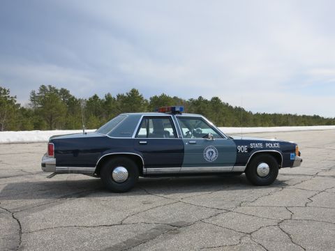 1991 Ford LTD Crown Victoria - Side profile of a two-tone blue Massachusetts State Police cruiser numbered 90E in an open paved lot.