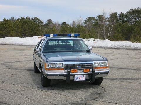 1991 Ford LTD Crown Victoria - Front view of a blue Ford police sedan with a push bumper, roof lightbar and a Massachusetts plate reading 90 E.
