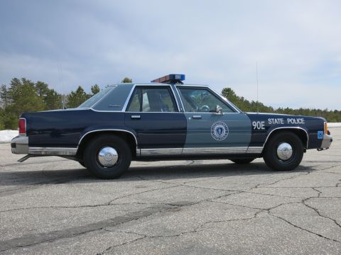 1991 Ford LTD Crown Victoria - Full side profile of a two-tone blue Massachusetts State Police sedan marked 90E parked on cracked asphalt.