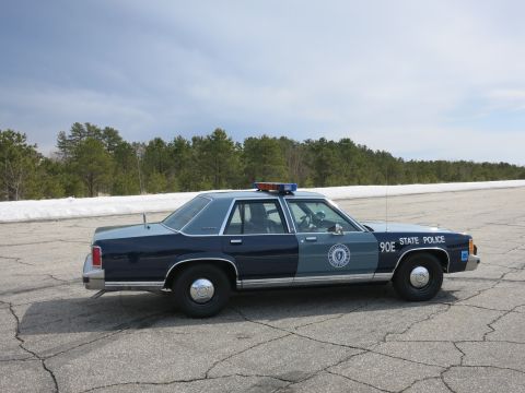 1991 Ford LTD Crown Victoria - blue Massachusetts State Police cruiser marked 90E shown in profile against pine trees and snow.