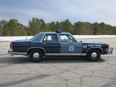 1991 Ford LTD Crown Victoria - Side view of a two-tone blue Massachusetts State Police sedan numbered 90E on a paved lot with pines behind.
