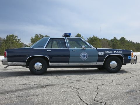 1991 Ford LTD Crown Victoria - Profile of a two-tone blue Massachusetts State Police sedan marked 90E parked on cracked asphalt under a cloudy sky.