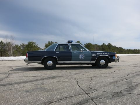 1991 Ford LTD Crown Victoria - blue Massachusetts State Police cruiser numbered 90E shown in profile in a wide paved lot with snow at the edges.