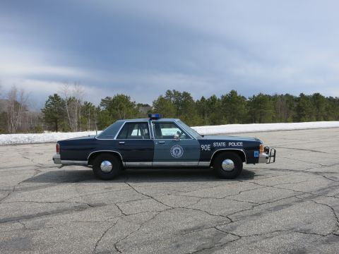 1991 Ford LTD Crown Victoria - Side view of a two-tone blue Massachusetts State Police sedan marked 90E with a front push bumper under cloudy skies.