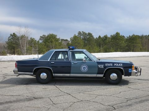 1991 Ford LTD Crown Victoria - blue Massachusetts State Police cruiser numbered 90E parked in profile on cracked pavement with pine trees behind.