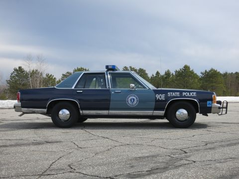 1991 Ford LTD Crown Victoria - Profile of a two-tone blue Massachusetts State Police sedan marked 90E parked on cracked asphalt under a gray sky.