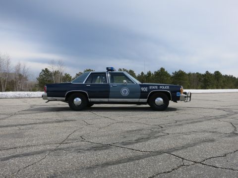1991 Ford LTD Crown Victoria - blue Massachusetts State Police cruiser numbered 90E with a push bumper, shown in profile in a wide paved lot.