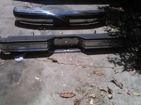 1992 Ford Bronco - Two chrome bumpers are shown, one with a black plastic step pad, lying on a concrete surface.