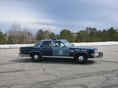 1991 Ford LTD Crown Victoria - Profile of a two-tone blue Massachusetts State Police sedan marked 90E parked on pavement with bare trees behind.