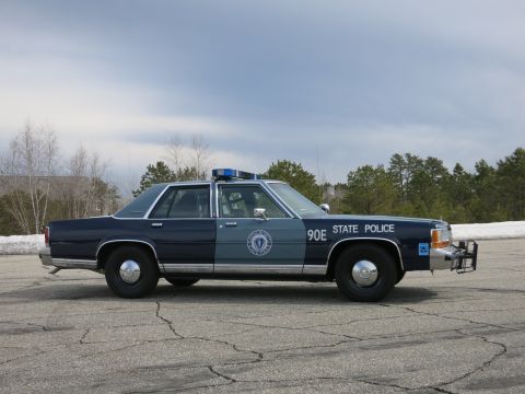 1991 Ford LTD Crown Victoria - blue Massachusetts State Police cruiser numbered 90E with a front push bumper shown in profile under a cloudy sky.