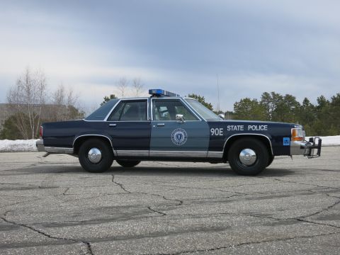 1991 Ford LTD Crown Victoria - Side view of a two-tone blue Massachusetts State Police sedan marked 90E with a push bumper near snowbanks and pines.