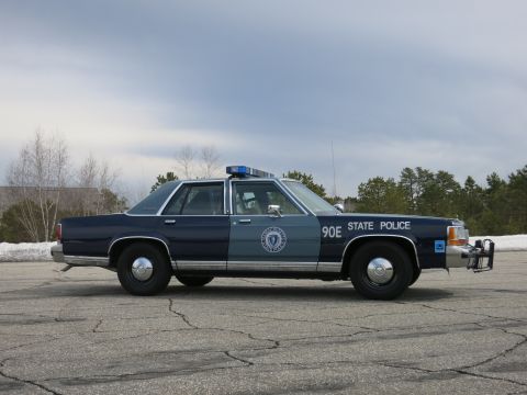 1991 Ford LTD Crown Victoria - blue Massachusetts State Police cruiser numbered 90E with a front push bumper shown in profile in a paved lot.