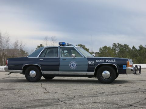 1991 Ford LTD Crown Victoria - Profile of a two-tone blue Massachusetts State Police sedan marked 90E parked on cracked asphalt with snow at the edges.