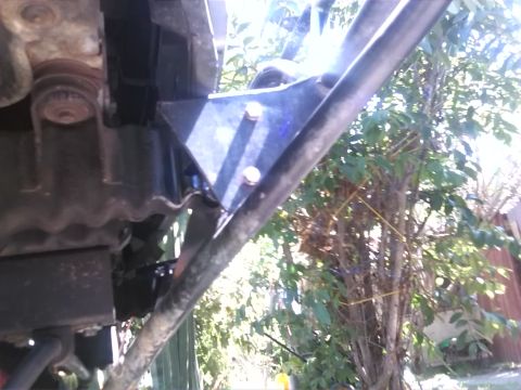1992 Ford Bronco - Detail of a vehicle's frame, showing a blue bracket attached with bolts. The surrounding foliage suggests an outdoor setting.