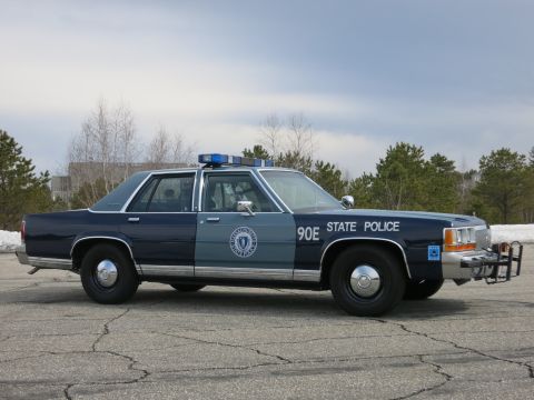 1991 Ford LTD Crown Victoria - two-tone dark and light blue State Police sedan marked 90E sits in profile on a cracked asphalt lot with a roof lightbar and front push bar.