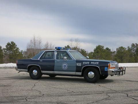 1991 Ford LTD Crown Victoria - two-tone blue State Police cruiser marked 90E stands broadside on a paved lot beneath a cloudy sky, fronted by trees.