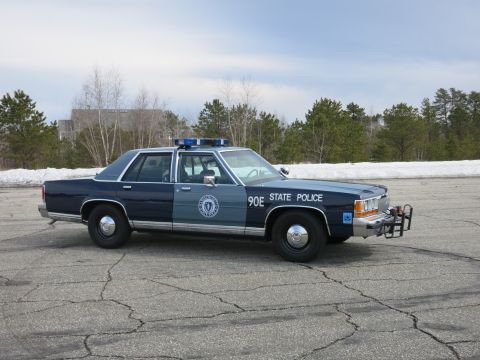1991 Ford LTD Crown Victoria - dark and light blue State Police sedan marked 90E shown front three-quarter with its blue lightbar and chrome push bumper on a cracked lot.