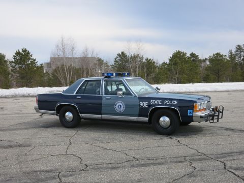 1991 Ford LTD Crown Victoria - two-tone blue State Police cruiser marked 90E poses at a front three-quarter angle on asphalt with melting snow and evergreens behind.