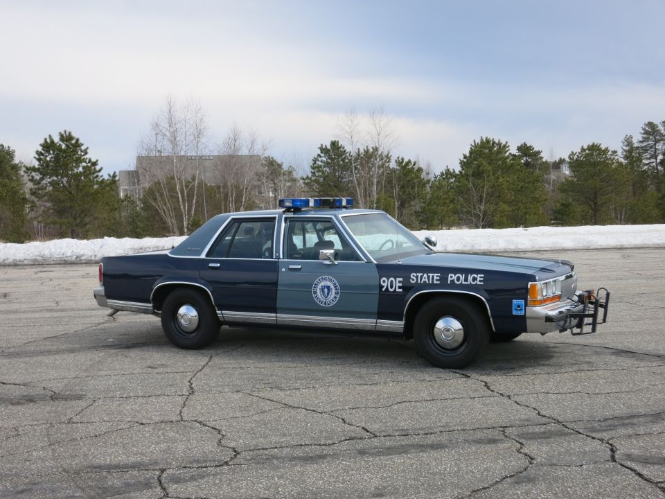 Blue State Police sedan, passenger front three-quarter, snow banks behind