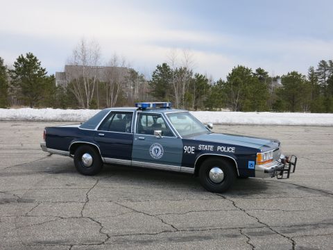 1991 Ford LTD Crown Victoria - dark and light blue State Police sedan marked 90E sits at a three-quarter angle on cracked asphalt with bare trees and snow behind.