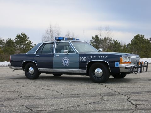 1991 Ford LTD Crown Victoria - two-tone blue State Police cruiser marked 90E shown front three-quarter, its lightbar and chrome push bumper catching grey daylight.