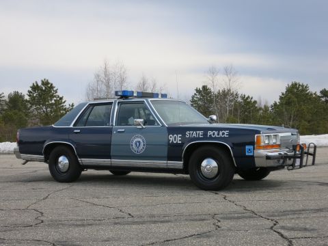1991 Ford LTD Crown Victoria - dark and light blue State Police sedan marked 90E stands at a three-quarter angle on a paved lot beneath an overcast sky.