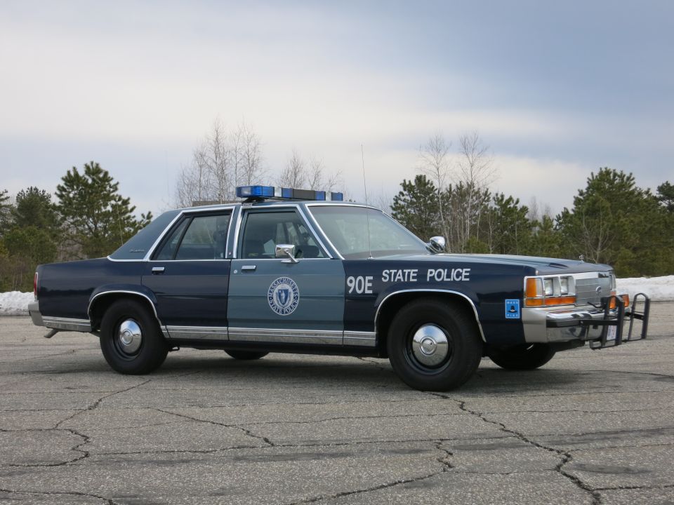 Two-tone blue State Police sedan, passenger three-quarter under cloudy sky